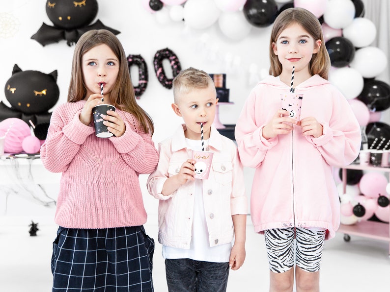 May include: Three children at a Halloween party, each holding a cup with a straw. The children are wearing pink clothing. The background includes black and white balloons and Halloween decorations. One child wears a pink sweater, another a pink jacket, and the third a pink zip-up hoodie.