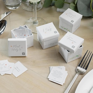 May include: White boxes with floral designs and the text "Shout Out!" "I Spy" "Trivia" and "Most Likely To" on them. Each box contains small cards with wedding-themed questions and games.