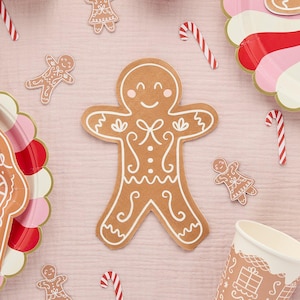 May include: A selection of gingerbread-themed party decorations. Features a large gingerbread man cutout, smaller gingerbread people, candy canes, and plates with pink and red details. The items are arranged on a pale pink surface.