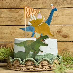 Könnte beinhalten: Ein dekorierter Kuchen mit Dinosaurier-Toppern. Der Kuchen hat eine weiße Frosting-Basis mit grünen und braunen dekorativen Elementen. Dinosaurier-Ausschnitte umfassen einen grünen T-Rex, einen gelben Triceratops und einen blauen Brontosaurus, alle mit Partyhüten. Eine Flagge liest "ROAR!"