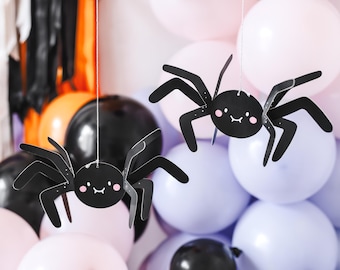 May include: Two black paper spider decorations with cute faces hanging from strings against a backdrop of pastel colored balloons.