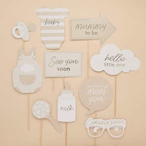 May include: A selection of baby shower photo booth props in beige and white. Props include a dummy, babygrow, dungarees, a sign saying "See you soon", a milk bottle, a rattle, a speech bubble, and glasses that say "Coming soon".