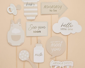 May include: A selection of baby shower photo booth props in beige and white. Props include a dummy, babygrow, dungarees, a sign saying "See you soon", a milk bottle, a rattle, a speech bubble, and glasses that say "Coming soon".