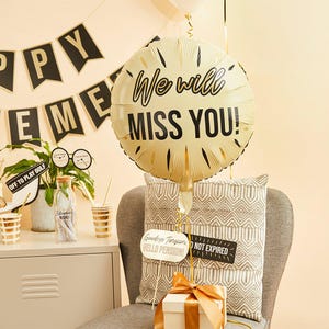 Puede incluir: Un globo dorado con el texto "We will miss you!" en letras negras. El globo está atado a una cuerda y está sentado en una silla con un cojín. También hay dos letreros pequeños con el texto "Goodbye Tension Hello Pension!" y "Not Expired" en la silla.