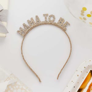 May include: A gold glitter headband with the words "MAMA TO BE" in capital letters. The headband is a party accessory, likely for a baby shower or gender reveal celebration. The headband is on a white surface.