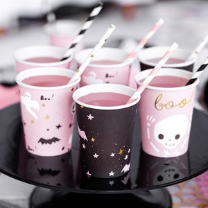 May include: Pink and black Halloween-themed paper cups filled with pink liquid, each with a straw. The cups feature spooky designs like ghosts, bats, stars, and the word "boo". They are arranged on a black cake stand.