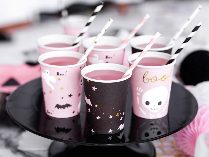 May include: Pink and black Halloween-themed paper cups filled with pink liquid, each with a straw. The cups feature spooky designs like ghosts, bats, stars, and the word "boo". They are arranged on a black cake stand.
