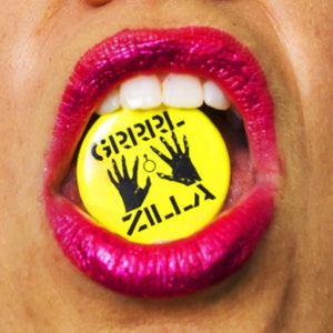 Puede incluir: Primer plano de una boca con lápiz labial rosa brillante que sostiene un botón amarillo. El botón tiene texto negro y dos siluetas de manos negras. El texto en el botón dice "ZILLA".
