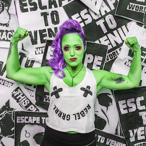 Puede incluir: Una mujer con piel verde y cabello morado está flexionando sus bíceps. Lleva un top blanco sin mangas con el texto "Rebel Grrrl" y pantalones negros. El fondo es un collage de carteles en blanco y negro con el texto "Escape to Venus" repetido.