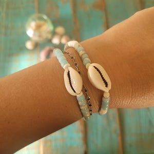 May include: Three beaded bracelets with a beach theme. Two bracelets feature light blue and white beads, with a large white seashell as a focal point. A delicate silver chain bracelet is also included. The bracelets are displayed on a turquoise surface.