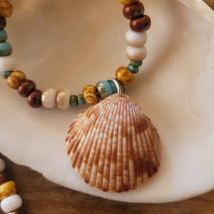 May include: A beaded necklace featuring a large, textured seashell pendant. The necklace has a strand of colorful beads in shades of brown, white, yellow, and turquoise. The seashell is a light brown color with vertical lines.