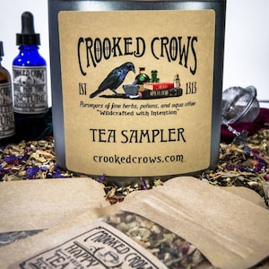 May include: A silver tin with a brown label that reads "CROOKED CROWS EST 1313 Purveyors of fine herbs, potions, and aqua vitae "Wildcrafted with Intention" TEA SAMPLER crookedcrows.com". The label features a black raven perched on a mortar and pestle. The tin is surrounded by small brown paper bags filled with loose leaf tea.
