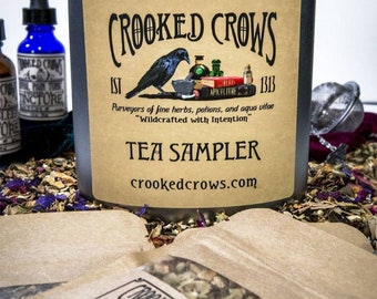 Peut inclure: Une boîte en étain argentée avec une étiquette brune qui indique "CROOKED CROWS EST 1313 Purveyors of fine herbs, potions, and aqua vitae "Wildcrafted with Intention" TEA SAMPLER crookedcrows.com". L'étiquette représente un corbeau noir perché sur un mortier et un pilon. La boîte est entourée de petits sachets en papier brun remplis de thé en feuilles.