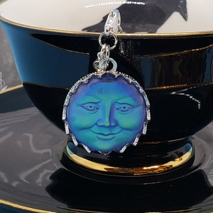 May include: A silver charm with a blue and green iridescent moon face, hanging from a silver chain. The charm is in a black teacup with a gold rim, on a black saucer with a gold rim. The text "CROOKED CROWS" is visible in the background.