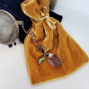 May include: A small, gold velvet pouch with a drawstring closure. The pouch is attached to a silver chain with a small silver charm in the shape of a crescent moon and a purple crystal. The pouch is sitting on a white surface.