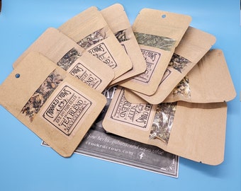 May include: Several small brown paper pouches with clear windows, filled with dried herbs, are arranged on a blue background. The pouches have labels with text that reads "Crooked Crows Tea Blend". The image suggests a tea product.