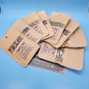May include: Several small brown paper pouches with clear windows, filled with dried herbs, are arranged on a blue background. The pouches have labels with text that reads "Crooked Crows Tea Blend". The image suggests a tea product.