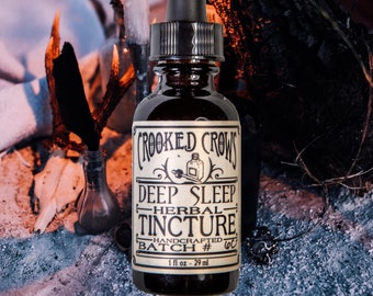May include: A dark brown glass bottle with a dropper top and a black label that reads "Crooked Crows Deep Sleep Herbal Tincture Handcrafted Batch # 60 1 fl oz - 29 ml". The bottle is sitting on a dark, textured surface with a candle in the background.