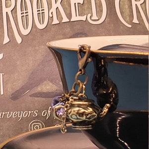 May include: A gold teapot charm with purple glass beads hanging from a black chain. The charm is hanging from the rim of a black teacup.