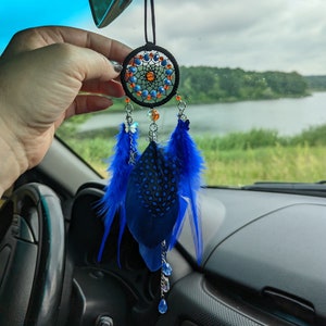 以下が含まれることがあります： 黒い円形のフレームに青とオレンジのビーズをあしらった手作りのドリームキャッチャー。鮮やかな青い羽根とぶら下がっているチャームが特徴で、黒いコードから吊り下げられています。ドリームキャッチャーは車内に吊るすように設計されています。