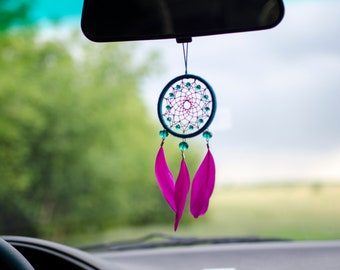 May include: A dreamcatcher hanging from a car's rearview mirror. The dreamcatcher is circular, with a web design in the center and adorned with turquoise beads and three bright pink feathers. The background shows a blurred green landscape.