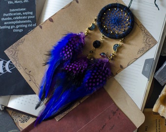 May include: A blue and purple dreamcatcher with feathers hanging from the bottom. The dreamcatcher is made of black thread and has a beaded design. The dreamcatcher is hanging from a notebook.