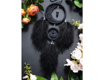 May include: Black dreamcatcher with a crescent moon and crystal charm, adorned with black feathers. The dreamcatcher is hanging against a dark background with pink flowers.