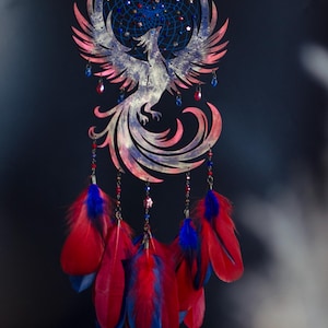 May include: A handcrafted dreamcatcher featuring a phoenix design. The dreamcatcher has a blue and red color scheme, with red and blue feathers hanging below. The phoenix is detailed with pink and white accents.