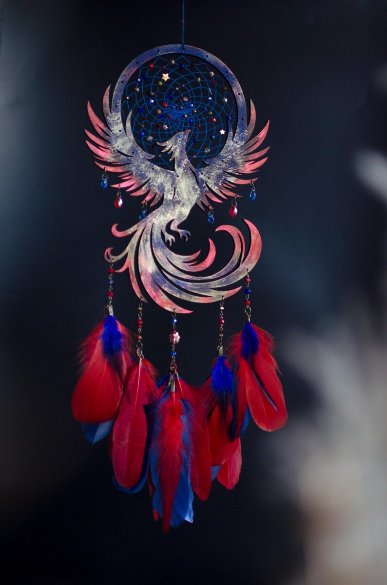 May include: A handcrafted dreamcatcher featuring a phoenix design. The dreamcatcher has a blue and red color scheme, with red and blue feathers hanging below. The phoenix is detailed with pink and white accents.