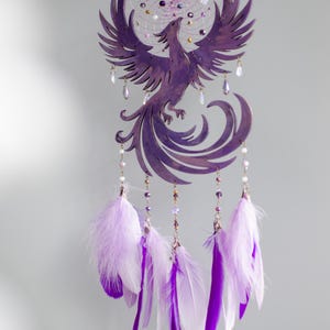 May include: Purple dreamcatcher featuring a phoenix design. The circular frame holds a web with beads and crystals. Below, the phoenix is crafted with outstretched wings. Hanging below are strands with beads and white and purple feathers.