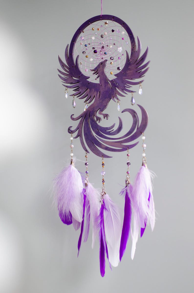 May include: Purple dreamcatcher featuring a phoenix design. The circular frame holds a web with beads and crystals. Below, the phoenix is crafted with outstretched wings. Hanging below are strands with beads and white and purple feathers.