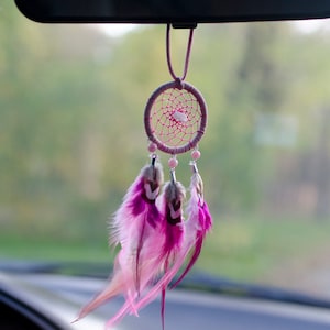 May include: A pink and white dreamcatcher with feathers hanging from a rearview mirror in a car.