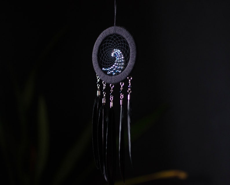 May include: A black dreamcatcher with a beaded design in the shape of a wave. The dreamcatcher has black feathers hanging from it.