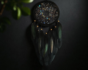 May include: A black dreamcatcher with a circular woven web, adorned with small, colorful beads and lights. Below, black feathers hang from the bottom. The dreamcatcher is suspended against a dark background, creating a striking contrast.