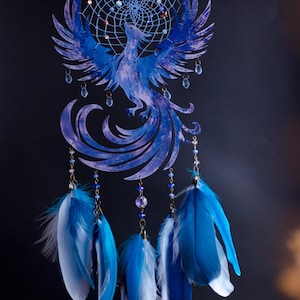 May include: A blue dreamcatcher featuring a phoenix design. The circular frame has a webbed center with beads. The phoenix is a detailed cutout with outstretched wings. Strands of beads and blue and white feathers hang below.