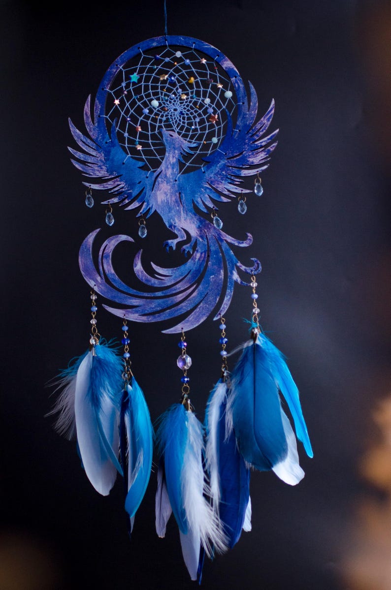 May include: A blue dreamcatcher featuring a phoenix design. The circular frame has a webbed center with beads. The phoenix is a detailed cutout with outstretched wings. Strands of beads and blue and white feathers hang below.