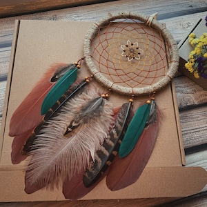 May include: A brown and beige dreamcatcher with a woven net and feathers. The feathers are brown, beige, green, and black. The dreamcatcher is hanging from a brown box.
