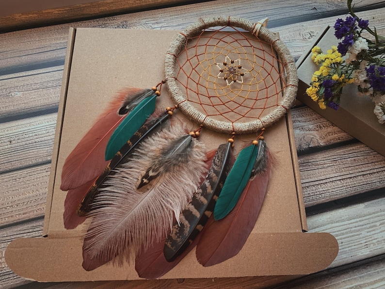 May include: A brown and beige dreamcatcher with a woven net and feathers. The feathers are brown, beige, green, and black. The dreamcatcher is hanging from a brown box.