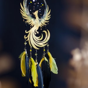 May include: A golden phoenix dreamcatcher with a black background. The phoenix is intricately cut with outstretched wings. Below, yellow and black feathers hang from the dreamcatcher, adding a touch of color and movement to the design.