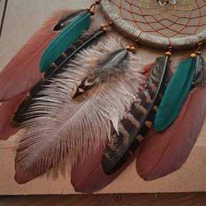 May include: A dream catcher with brown and white feathers, including a long white feather with brown tips, and two teal feathers. The dream catcher has a brown leather cord and beads.