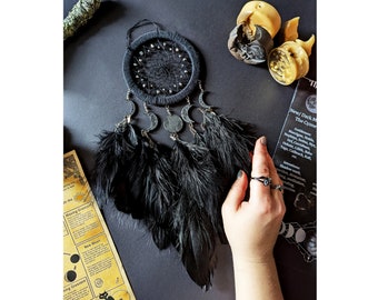 May include: Black dreamcatcher with feathers and moon charms. The dreamcatcher is hanging from a black cord. The background is a dark gray surface.