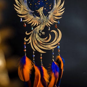 Handcrafted phoenix-shaped dreamcatcher with a deep blue woven web, orange and blue feathers, star beads, and crystal drops. Symbolizes rebirth and strength. Perfect for fantasy-themed or boho-style home decor.