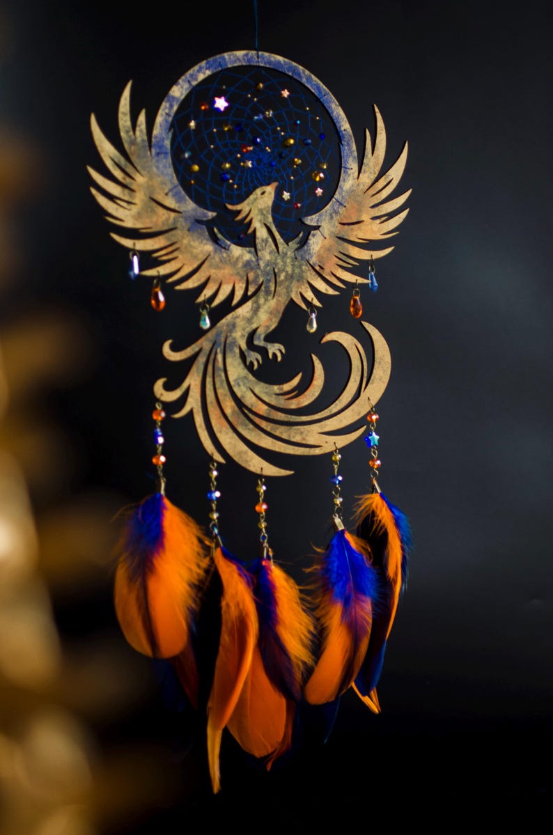 Handcrafted phoenix-shaped dreamcatcher with a deep blue woven web, orange and blue feathers, star beads, and crystal drops. Symbolizes rebirth and strength. Perfect for fantasy-themed or boho-style home decor.