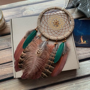 May include: A brown and beige dreamcatcher with green feathers and brown and white feathers. The dreamcatcher is hanging from a brown box with a white card on the side.
