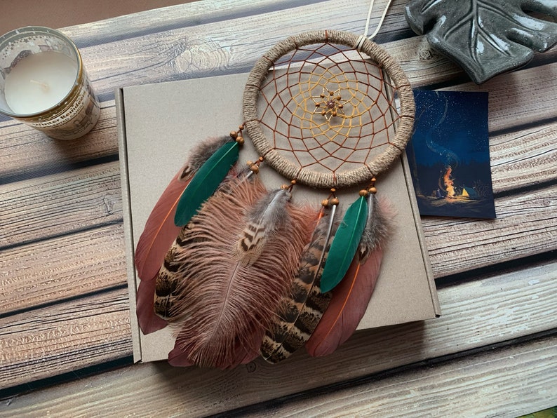 May include: A brown and beige dreamcatcher with green feathers and brown and white feathers. The dreamcatcher is hanging from a brown box with a white card on the side.