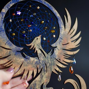 Handcrafted phoenix-shaped dreamcatcher with a deep blue woven web, orange and blue feathers, star beads, and crystal drops. Symbolizes rebirth and strength. Perfect for fantasy-themed or boho-style home decor.