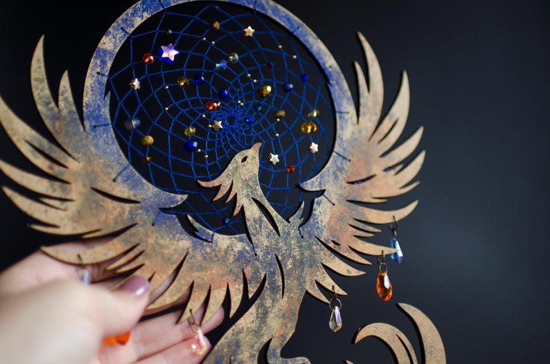 Handcrafted phoenix-shaped dreamcatcher with a deep blue woven web, orange and blue feathers, star beads, and crystal drops. Symbolizes rebirth and strength. Perfect for fantasy-themed or boho-style home decor.
