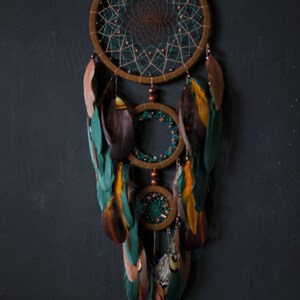 May include: A brown and green dreamcatcher with feathers. The dreamcatcher has three hoops and is decorated with beads. The feathers are a mix of brown, green, and orange.