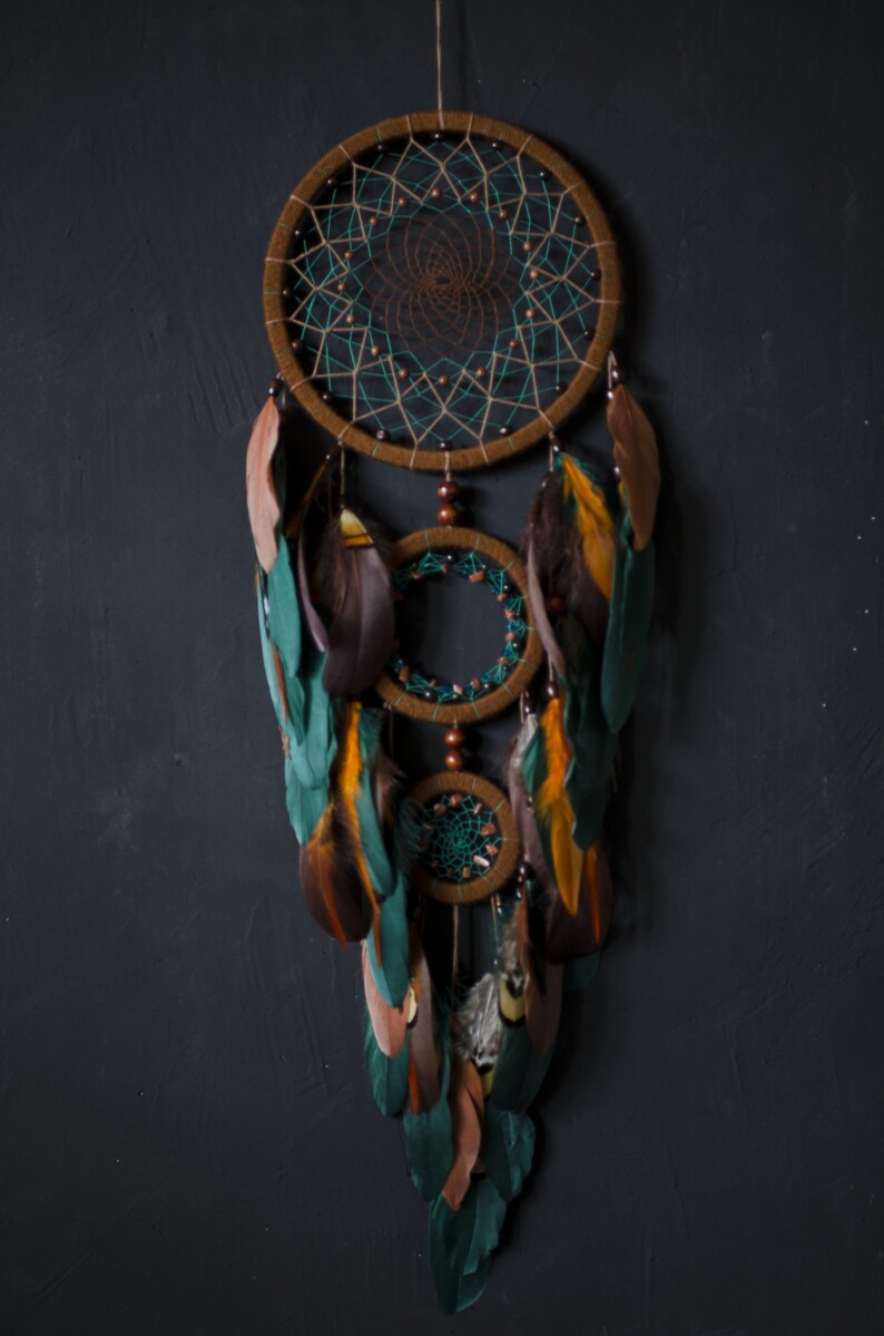 May include: A brown and green dreamcatcher with feathers. The dreamcatcher has three hoops and is decorated with beads. The feathers are a mix of brown, green, and orange.