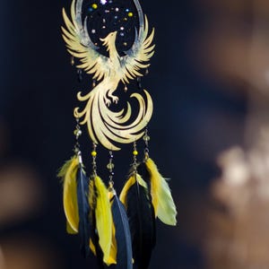 May include: A golden phoenix dreamcatcher with a black background. The phoenix is intricately cut with outstretched wings. Below, yellow and black feathers hang from beaded strands, adding a touch of color and movement to the design.